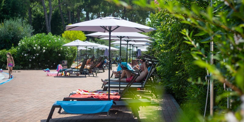 Lounge chairs with people relaxing under umbrellas beside a pool, surrounded by lush greenery at Del Sole