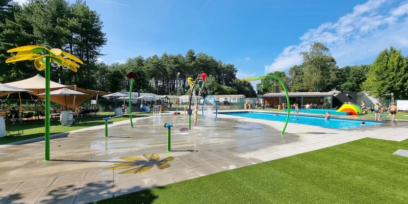 Colorful water park features fountains and playful structures near a swimming pool, surrounded by trees and green grass at Vakantiepark Breebos