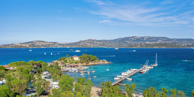 Boats are docked along a pier in clear blue water, surrounded by lush trees and distant mountains at Isuledda