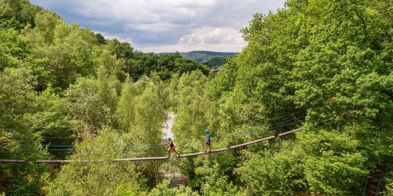 Two people, crossing a suspension bridge, surrounded by dense forest, and cloudy sky at Landal Village les Gottales
