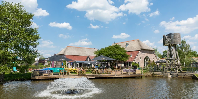 Fountain spouting water in pond with rustic building, patio umbrellas, and wooden tower on sunny day at Villapark de Weerribben