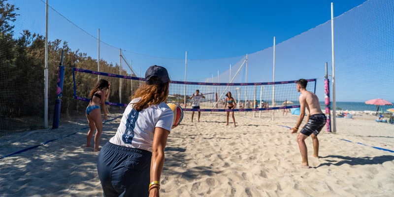 People play beach volleyball, setting and spiking, on a sandy court by the ocean under a clear blue sky at Stork Family Camping Village