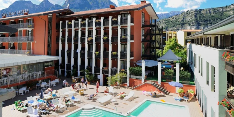 People relaxing on lounge chairs beside a swimming pool, surrounded by hotel buildings with mountains in the background at Holiday Park La Vela