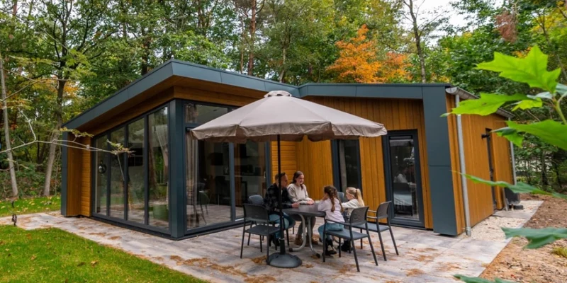 Modern cabin with large windows; family dining under patio umbrella; surrounded by lush, green forest at Europarcs de Wiltzangh
