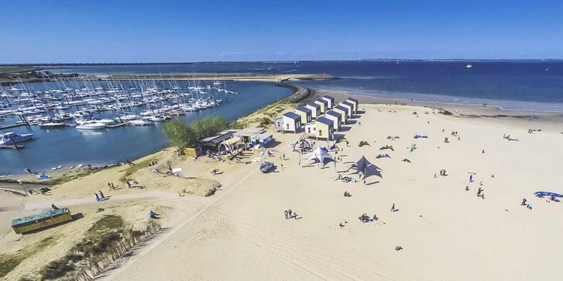 Beach-goers relaxing on sandy shore near white cabins and boats docked in a marina under clear blue sky at Strandpark Duynhille