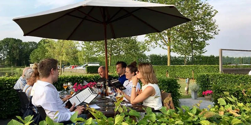 People dine under a large patio umbrella at a table surrounded by greenery and blooming flowers at Betuwe