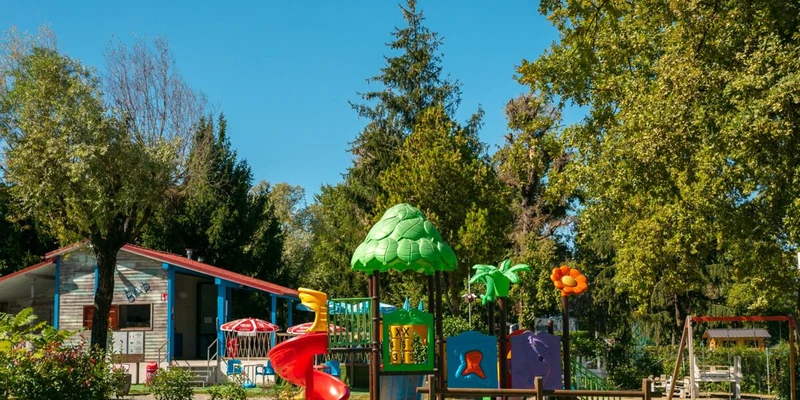 Colorful playground structures with slides and swings, adjacent to a small building amid lush trees at Lido Verbano