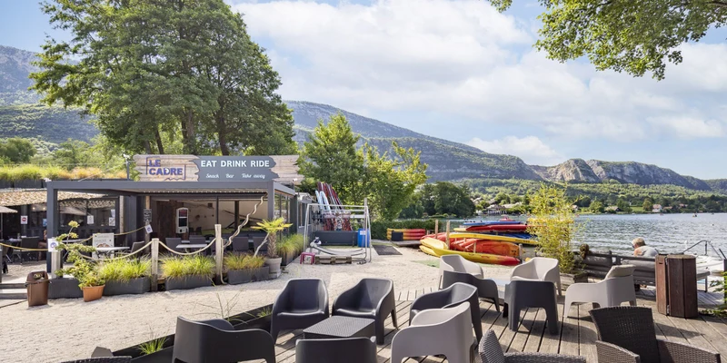 Outdoor café seating near a lake, surrounded by kayaks, trees, and mountains, under a partly cloudy sky at La Nublière