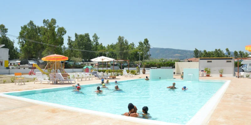 People swimming and relaxing in a sunlit outdoor pool with umbrellas and trees at Tindari Village