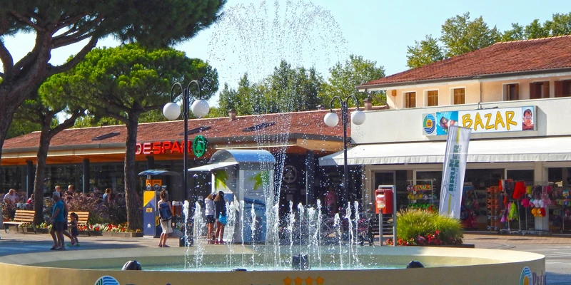 Fountain spraying water in front of shops surrounded by trees and people walking at Pra delle Torri
