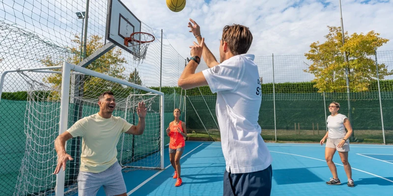 Four people playing basketball on a bright blue outdoor court, surrounded by netting and trees at Europa Camping Village