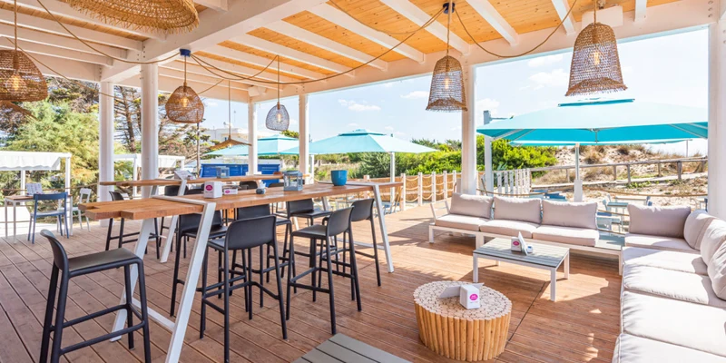 Outdoor seating area with high tables and lounge chairs on a wooden deck, surrounded by wicker lamps and umbrellas at Porto Cesareo Camping