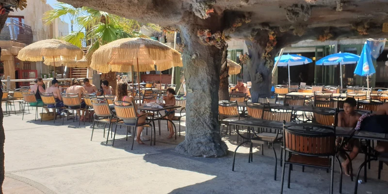 People sitting under shaded umbrellas, relaxing and dining in a sunlit outdoor café at Resort Cap Pirate