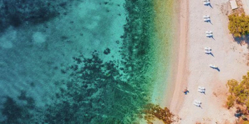 Aerial view of turquoise sea meeting sandy beach with scattered lounge chairs; trees border the shoreline at Boutique Camping Bunja