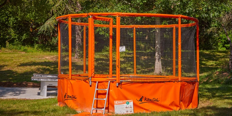Bright orange trampoline, surrounded by safety netting, with a small ladder, located in a grassy, wooded area at Le Chêne Vert