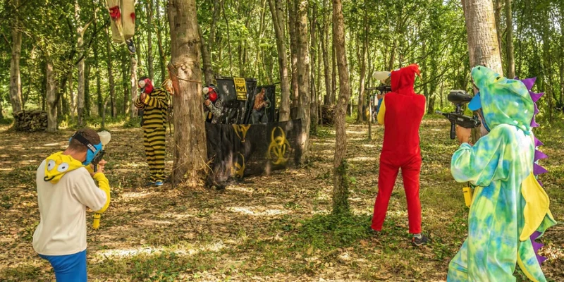 People in colorful costumes playing paintball in a forest clearing, surrounded by trees and makeshift barriers at Marvilla Parks Château la Forêt