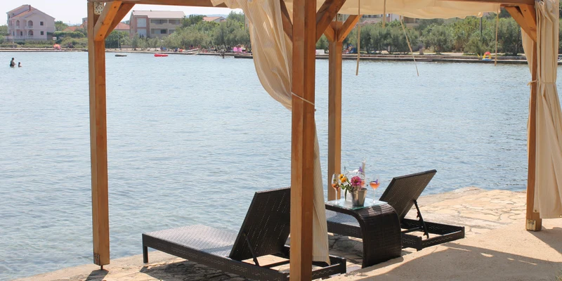 Two lounge chairs under a curtained pergola facing a calm waterfront; buildings visible in the background at Camping Taber Pašman