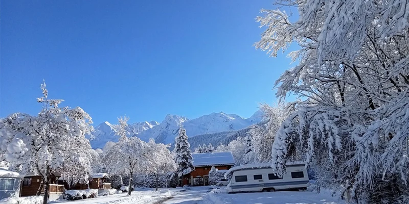 A caravan is parked on a snowy campsite path with snow-covered trees and mountains in the background at Clos du Pin