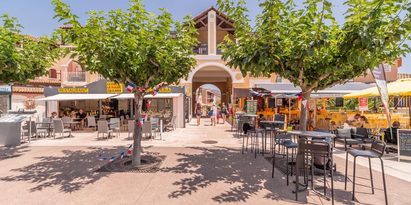 Restaurants and cafes line a sunny plaza with trees, umbrellas, and people walking under a building archway at Village Cap Esterel