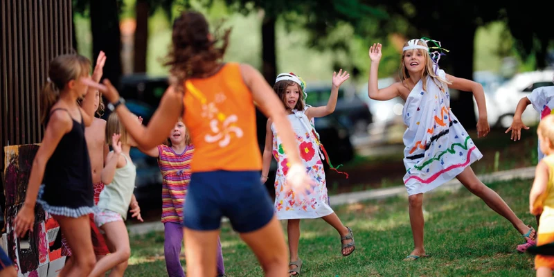 Children playing a group game outdoors led by an adult, with trees and cars in the background at Naturist Park Koversada
