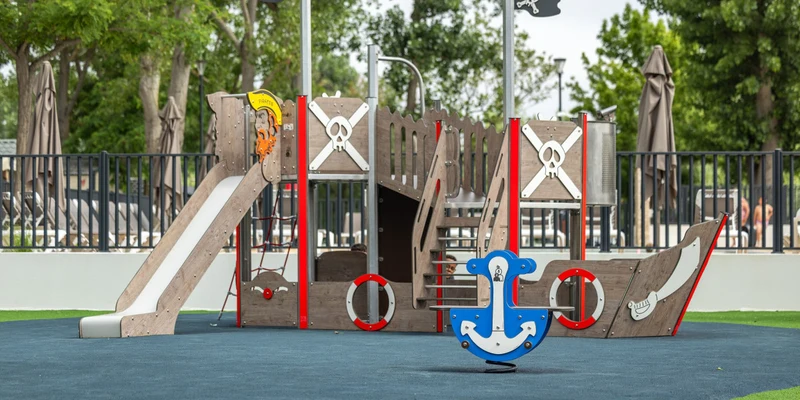 Pirate-themed playground structure features slide and climbing areas, surrounded by trees and fenced outdoor area at Marvilla Parks La Marine