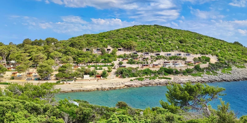 Hillside cabins overlook a turquoise bay, surrounded by lush trees under a partly cloudy sky at Poljana