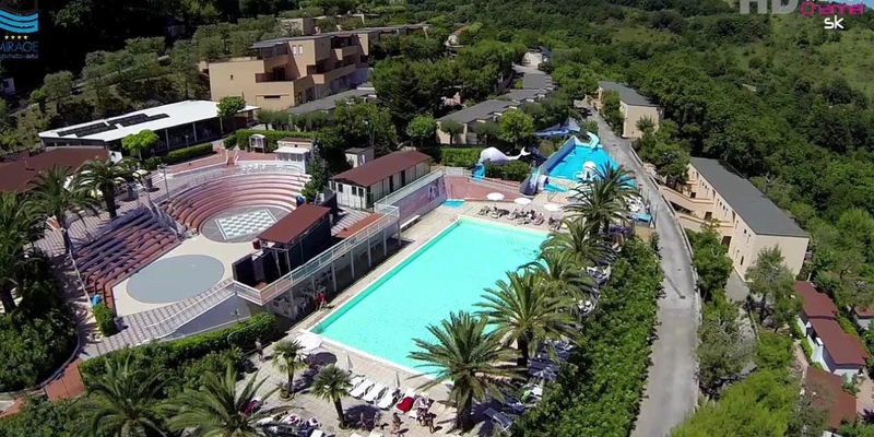 Swimming pool surrounded by deck chairs and palm trees, located near an amphitheater and buildings, amidst lush greenery at Camping Village Mirage