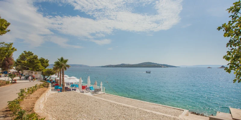 Shade umbrellas and lounge chairs line a beach-facing terrace beside a gravelly shore with trees and distant islands, at Amadria Park Camping Trogir