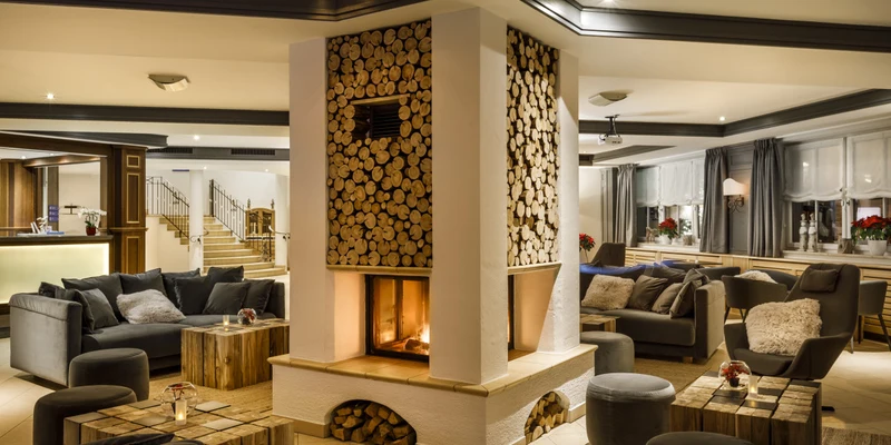 Central fireplace burning warmly, surrounded by cozy seating and wooden decor, within a spacious, elegantly furnished lounge at Valamar Obertauern Hotel