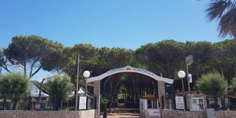 Entrance gate with stone pillars stands open, welcoming visitors into a forested camping area, under a clear blue sky at Camping Serenella Puglia