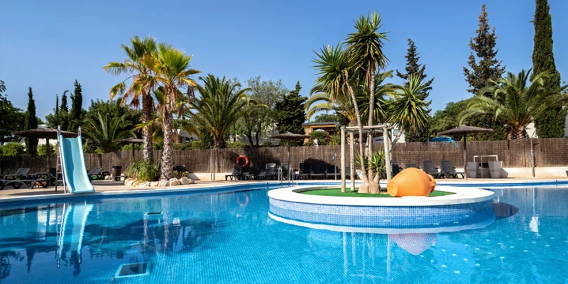 Swimming pool with a central island featuring palm trees, surrounded by lounge chairs and umbrellas under a clear blue sky at Orangeraie