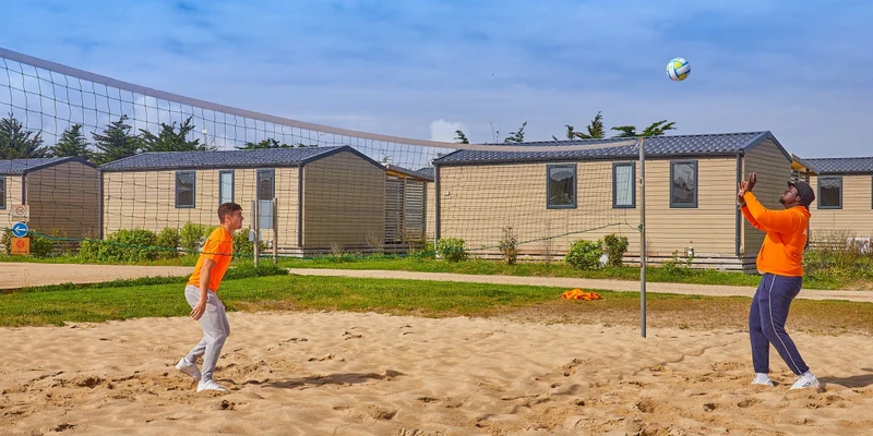 Two people play beach volleyball; mobile homes are visible in the background at Marvilla Parks Les Chardons Bleus de la Turballe