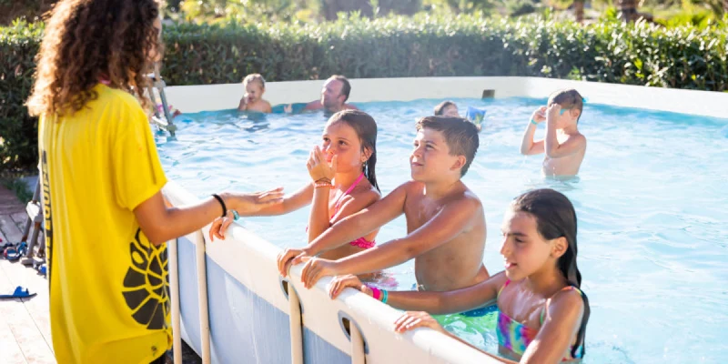 Children stand in a pool, talking to an instructor in a yellow shirt; surrounded by greenery at Triscina Mare