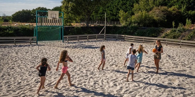Children playing with a ball on a sandy court surrounded by a wooden fence and trees at Les Brillas
