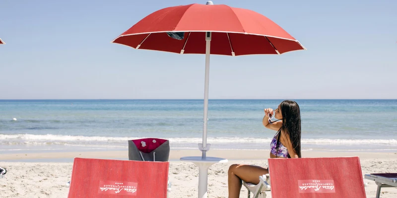 Woman sitting under a red umbrella, adjusting sunglasses on a sandy beach with the ocean in the background at Torre Rinalda