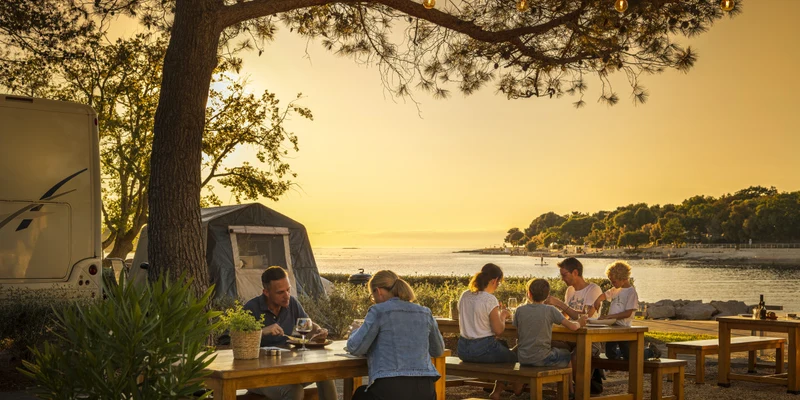 People dining outdoors on wooden tables, by a camper and a tent with a sunset view over the water at Valkanela