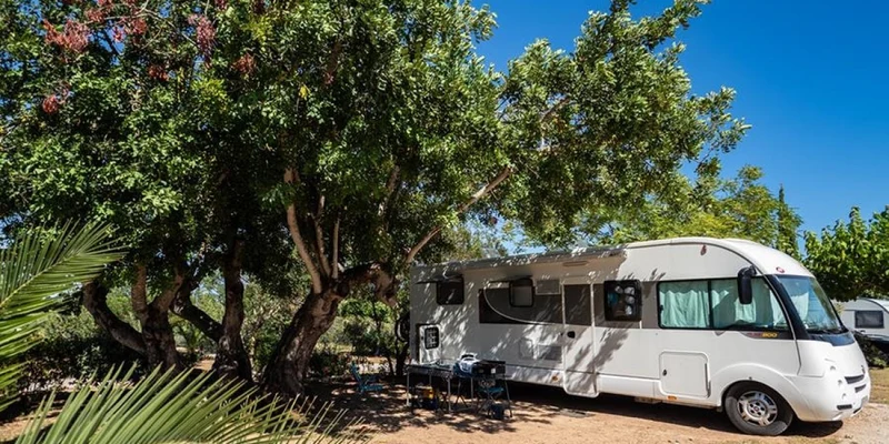 White RV parked under large shady trees, with outdoor furniture set up nearby at Orangeraie