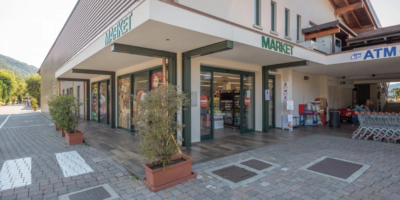 Market building with large windows and potted plants, adjacent to a parking area and surrounded by greenery at Isolino