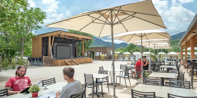 People sitting at outdoor tables under large umbrellas near a stage, surrounded by trees and mountains at Terra Verdon 