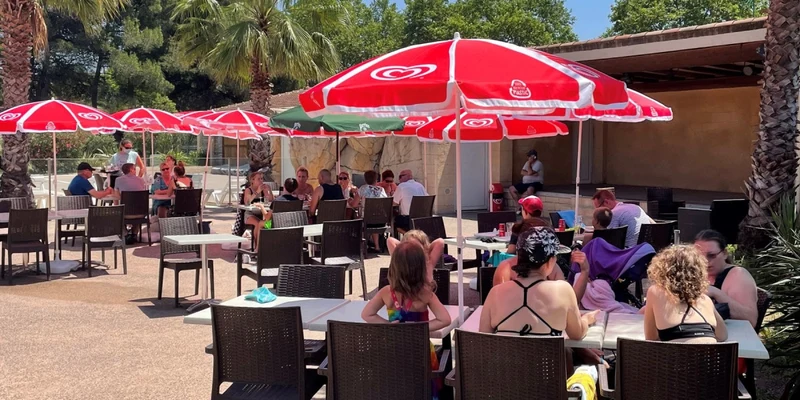 People seated under red umbrellas, eating and socializing in an open-air cafe surrounded by palm trees at La Prairie 