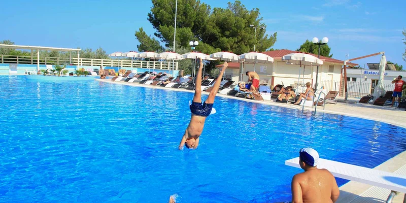 Child diving into a swimming pool while others relax on lounge chairs under umbrellas at Village Baia del Monaco