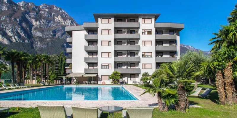 A multi-story building stands behind a large swimming pool surrounded by palm trees and mountains in the background at Residence Monica