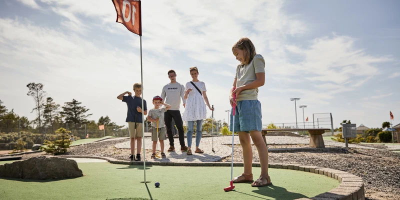 Child putting a golf ball on a miniature golf course while their family watches at Skiveren Camping