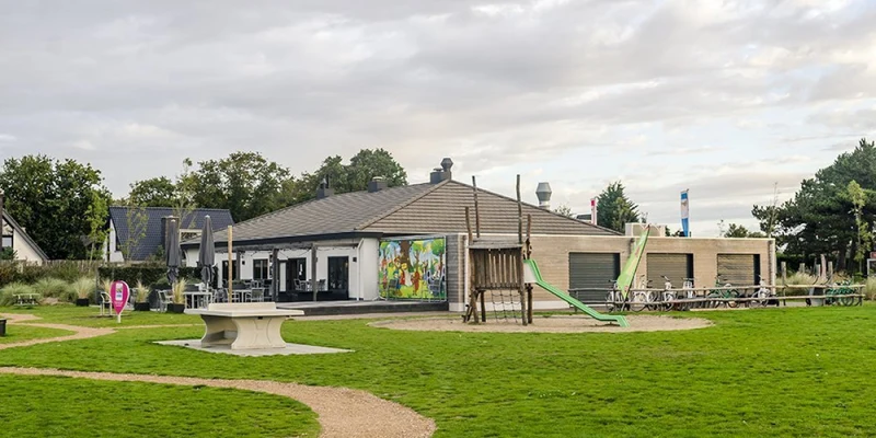 Playground slide descends from wooden structure, situated on grassy area near modern recreational building with picnic tables at Strandpark Duynhille