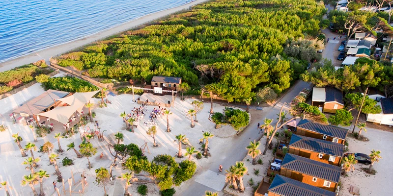 People gather on a sandy beach near wooden cabins and lush greenery, adjacent to a calm blue sea at Eurosurf