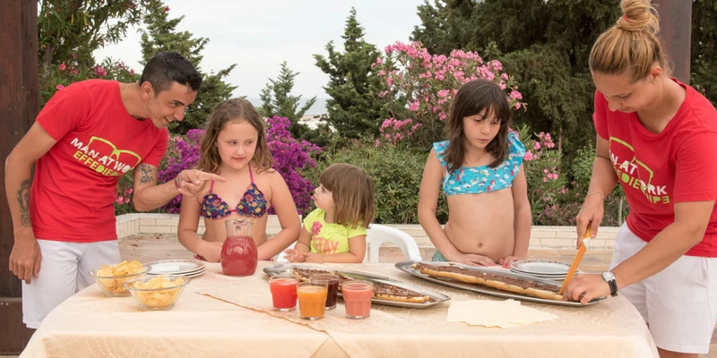 Two adults and three children preparing and serving food at a table outdoors, surrounded by blooming plants and trees at Villaggio Club Altalia