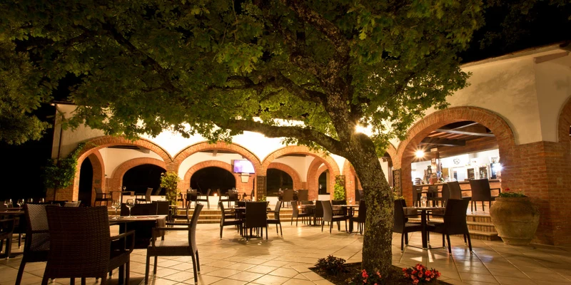 Outdoor dining area with wicker chairs and tables under a tree, in front of a building with brick arches at Luxor Chianti Glamping Village