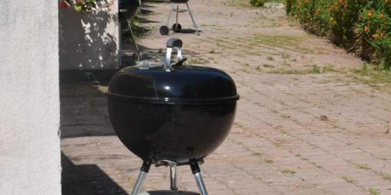 Black charcoal grills stand idle on sunlit pavement beside a lush garden at Camping & Residence Villa Teti