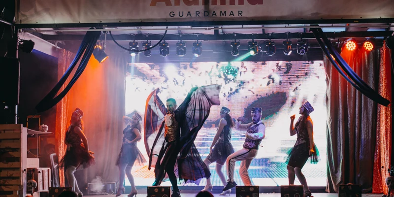 Performers dance energetically on a lit stage, surrounded by dark curtains and an audience at Alannia Guardamar