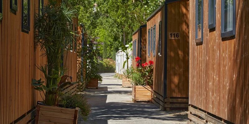 Cabins with wooden exteriors line a shaded path, adorned with vibrant potted plants at Marvilla Parks Verdon Parc